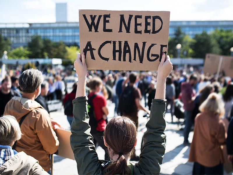 A lady standing in crowd by holding a bord of need a change