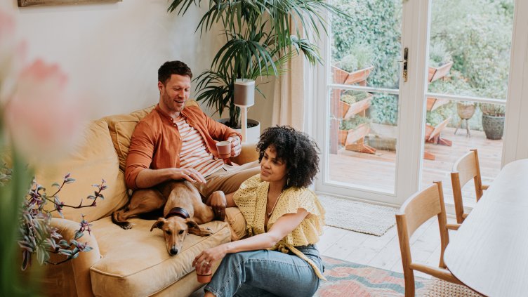 A beautiful young couple relax together in a stylish living room. The male sits on a soft, luxurious velvet yellow sofa as a lurcher dog lies curled up beside him. The female sits on a rug on the ground in front of her partner. They smile.